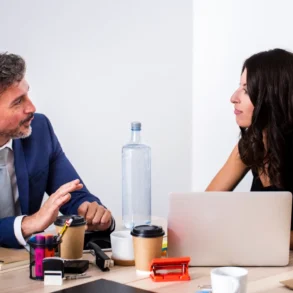 A man in a blue suit and a woman in a black top are in a focused discussion at a desk with laptops, coffee cups, and office supplies.
