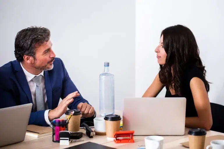 A man in a blue suit and a woman in a black top are in a focused discussion at a desk with laptops, coffee cups, and office supplies.