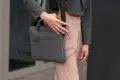 Person wearing beige pants and a gray blazer holding a gray fabric laptop bag with a shoulder strap and a hand on the strap.