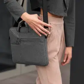 Person wearing beige pants and a gray blazer holding a gray fabric laptop bag with a shoulder strap and a hand on the strap.