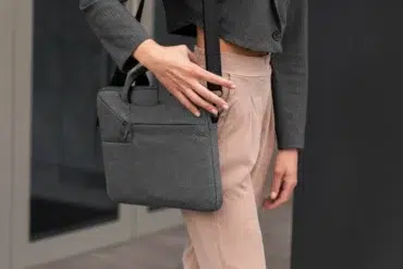 Person wearing beige pants and a gray blazer holding a gray fabric laptop bag with a shoulder strap and a hand on the strap.