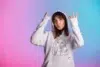 Young woman in a white hoodie adorned with a line drawing poses playfully against a pink and blue gradient background, expressing a cheerful vibe.
