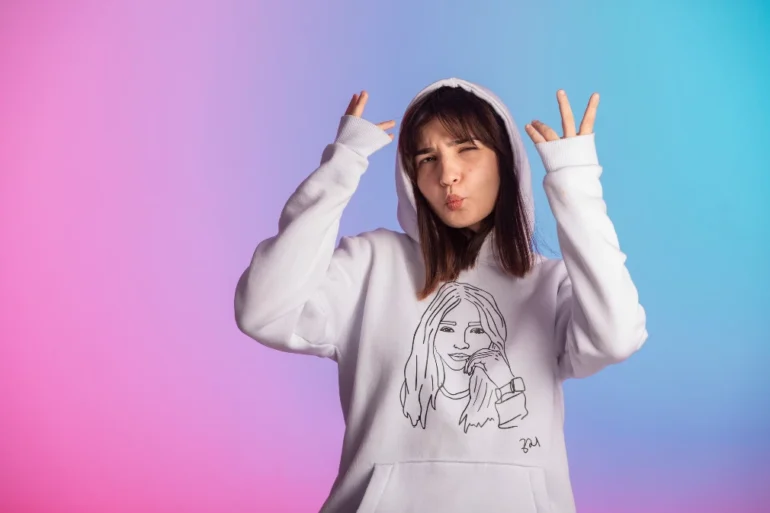 Young woman in a white hoodie adorned with a line drawing poses playfully against a pink and blue gradient background, expressing a cheerful vibe.