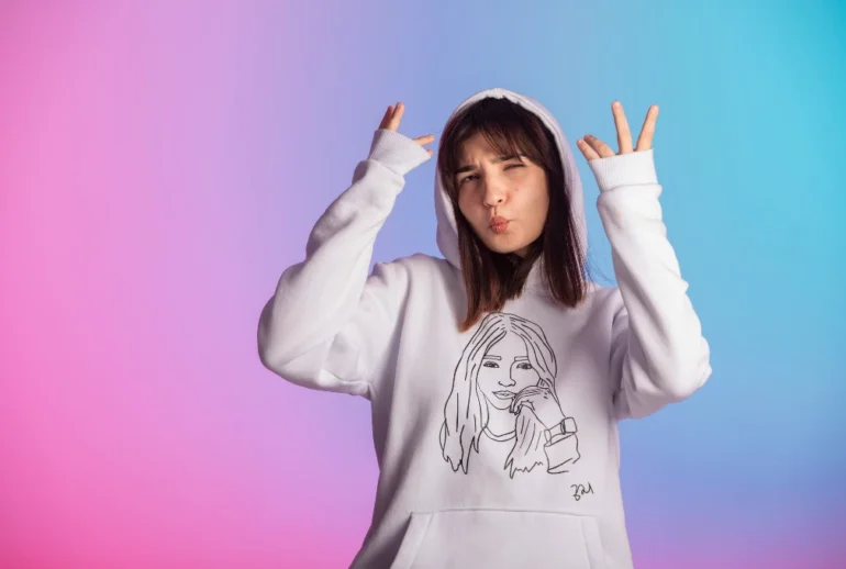 Young woman in a white hoodie adorned with a line drawing poses playfully against a pink and blue gradient background, expressing a cheerful vibe.