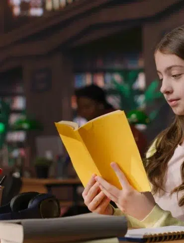 Young girl in a library reads a yellow book, illuminated by a green desk lamp. Her expression is focused. Background shows shelves filled with books.