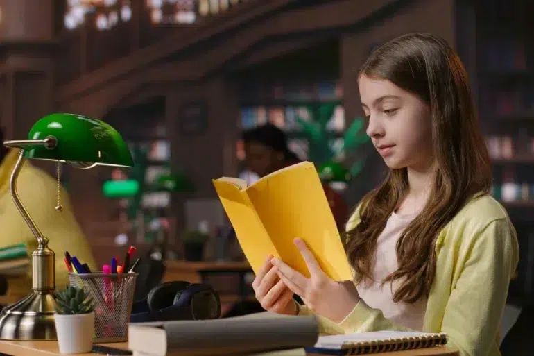 Young girl in a library reads a yellow book, illuminated by a green desk lamp. Her expression is focused. Background shows shelves filled with books.
