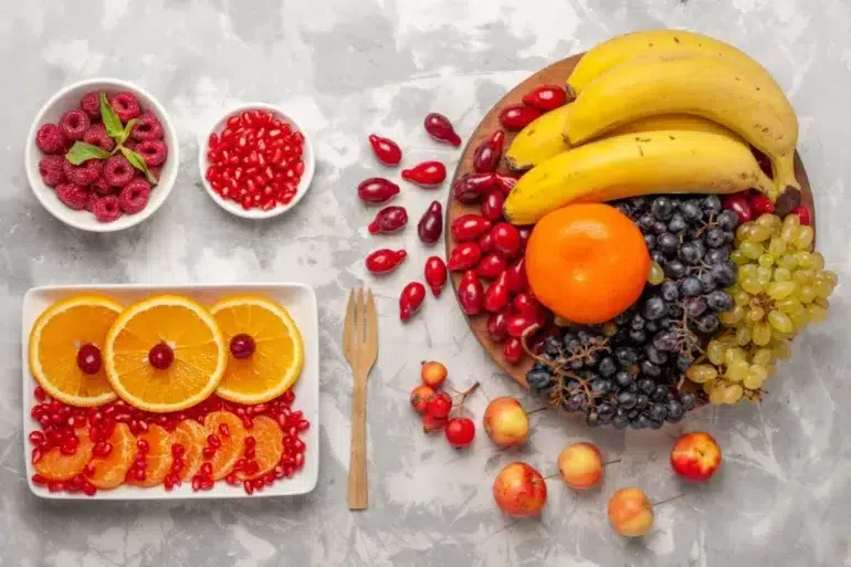 Assortment of colorful fruits on a marble surface. Bananas, orange, grapes, and apples on a wooden plate. Sliced oranges, raspberries, and pomegranate in bowls. Festive and fresh.