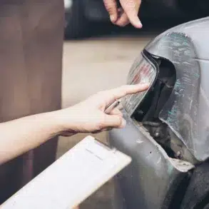 Two people inspect a damaged car bumper. One points at a dent near the headlight, holding a pen and clipboard, suggesting an accident report.