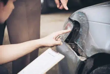Two people inspect a damaged car bumper. One points at a dent near the headlight, holding a pen and clipboard, suggesting an accident report.