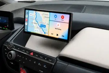 Car dashboard with a digital display showing navigation map and music controls. The map highlights a route near San Mateo. Buttons below are for climate control.