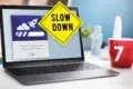 A laptop displays a progress bar with a rocket icon, overlaid by a yellow "Slow Down" sign. A red mug with "7" sits nearby, suggesting a cautionary tone.