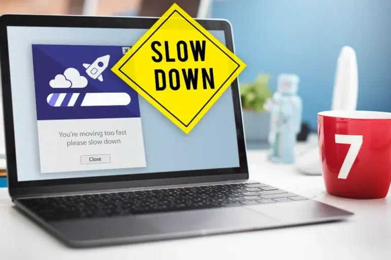 A laptop displays a progress bar with a rocket icon, overlaid by a yellow "Slow Down" sign. A red mug with "7" sits nearby, suggesting a cautionary tone.