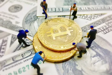 Figurines work on large stacked Bitcoin coins placed on US dollar bills. The scene conveys the concept of cryptocurrency mining and finance.