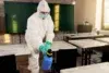 Person in full protective gear sanitizes a classroom with a spray bottle. Empty desks and a green chalkboard are visible, conveying a clean and safe environment.