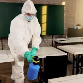 Person in full protective gear sanitizes a classroom with a spray bottle. Empty desks and a green chalkboard are visible, conveying a clean and safe environment.
