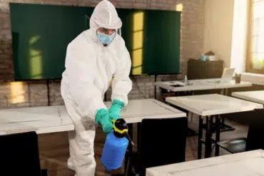Person in full protective gear sanitizes a classroom with a spray bottle. Empty desks and a green chalkboard are visible, conveying a clean and safe environment.