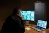 A person in a dim room monitors multiple CCTV feeds on a large screen and a laptop at a wooden desk, conveying focus and vigilance in security work.