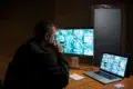 A person in a dim room monitors multiple CCTV feeds on a large screen and a laptop at a wooden desk, conveying focus and vigilance in security work.
