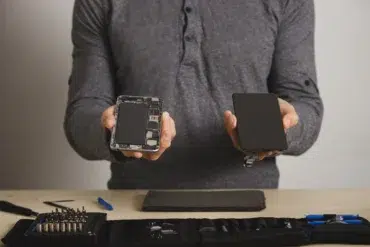 A person in a grey shirt holds a disassembled smartphone and a replacement screen, with repair tools on a table, conveying a tech repair theme.