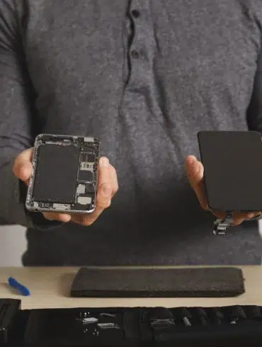 A person in a grey shirt holds a disassembled smartphone and a replacement screen, with repair tools on a table, conveying a tech repair theme.