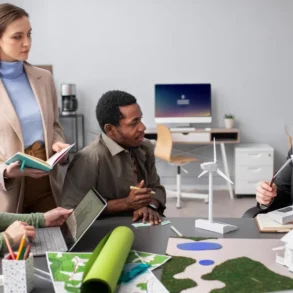 Four people discuss a green energy project in an office. They examine model wind turbines and building plans. The atmosphere is collaborative and focused.