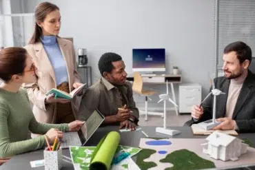 Four people discuss a green energy project in an office. They examine model wind turbines and building plans. The atmosphere is collaborative and focused.