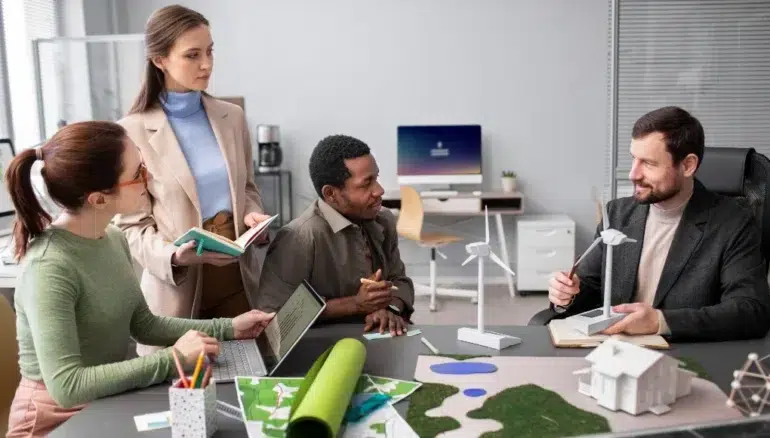 Four people discuss a green energy project in an office. They examine model wind turbines and building plans. The atmosphere is collaborative and focused.