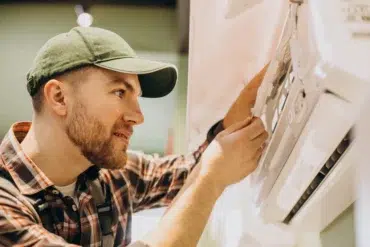 A man in a green cap and plaid shirt repairs an air conditioning unit with focus and concentration, highlighting a professional and skilled demeanor.