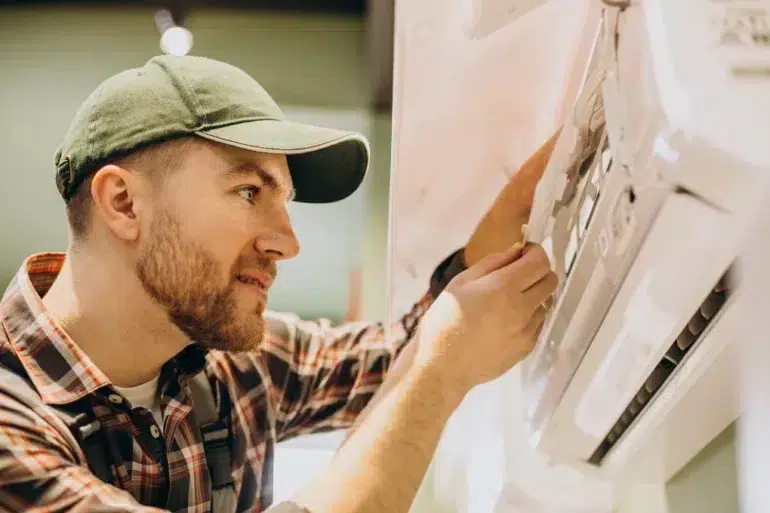 A man in a green cap and plaid shirt repairs an air conditioning unit with focus and concentration, highlighting a professional and skilled demeanor.