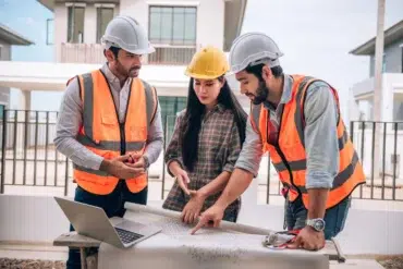 Three construction professionals in safety vests and helmets review architectural blueprints on a table. They display focus and collaboration.
