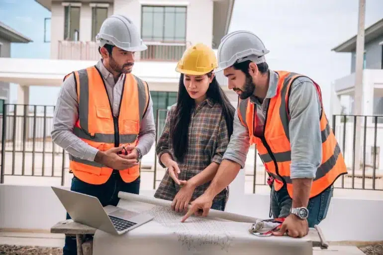 Three construction professionals in safety vests and helmets review architectural blueprints on a table. They display focus and collaboration.