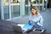 A woman in a blue shirt works outside on a wooden table, with a laptop, tablet, phone, and blue folder. She appears focused and thoughtful.