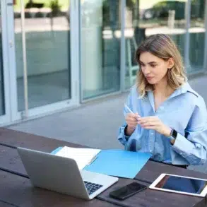 A woman in a blue shirt works outside on a wooden table, with a laptop, tablet, phone, and blue folder. She appears focused and thoughtful.