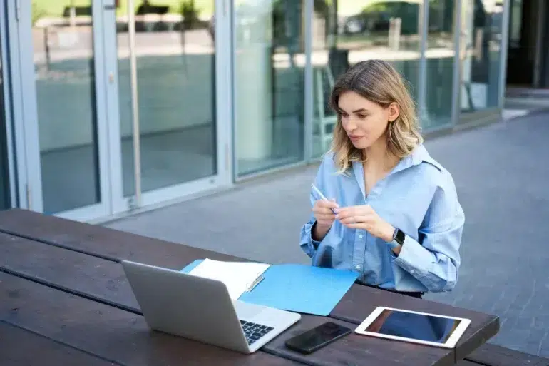 A woman in a blue shirt works outside on a wooden table, with a laptop, tablet, phone, and blue folder. She appears focused and thoughtful.
