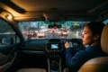 Inside a car during evening traffic, a woman drives attentively on a wet road. Dashboard lights glow as taillights illuminate the rainy scene outside.