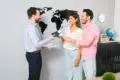 A couple smiles while receiving travel documents from a professional in an office with a world map on the wall, suggesting travel planning.