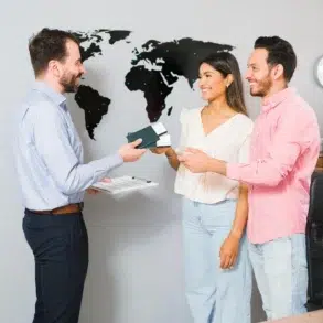 A couple smiles while receiving travel documents from a professional in an office with a world map on the wall, suggesting travel planning.