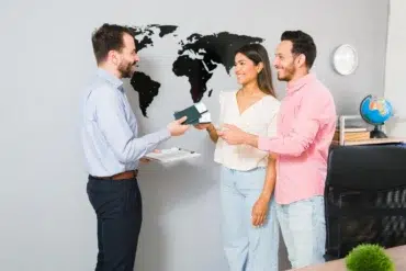 A couple smiles while receiving travel documents from a professional in an office with a world map on the wall, suggesting travel planning.