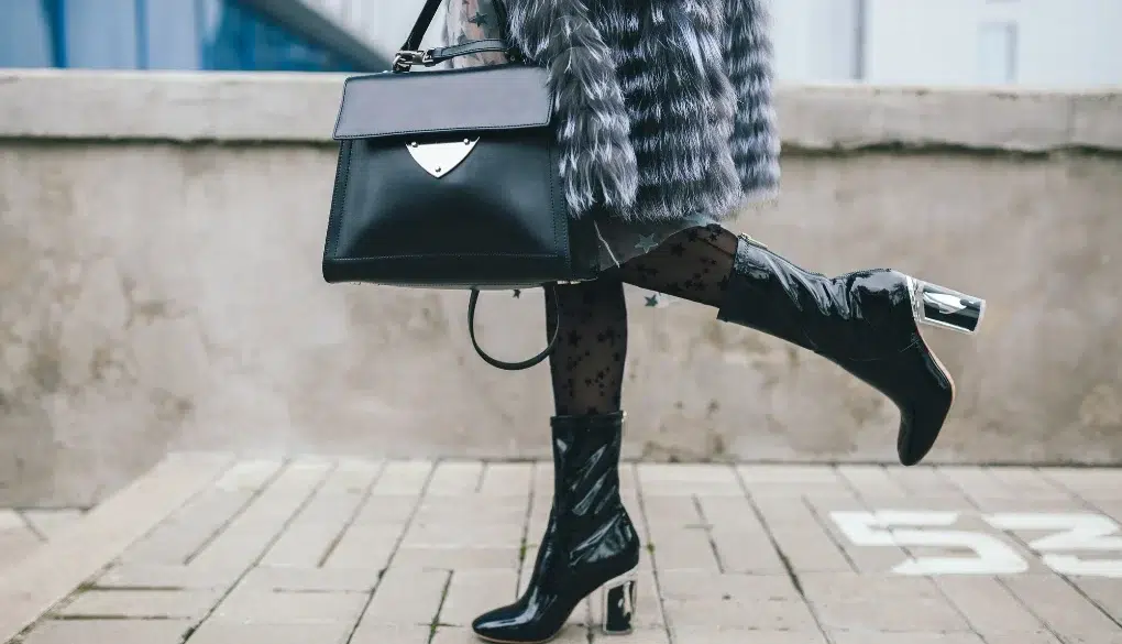Fashionable person in a fur coat carries a black handbag and wears sheer polka-dot tights with glossy black boots, exuding chic style.