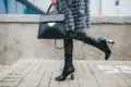 Fashionable person in a fur coat carries a black handbag and wears sheer polka-dot tights with glossy black boots, exuding chic style.