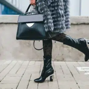 Fashionable person in a fur coat carries a black handbag and wears sheer polka-dot tights with glossy black boots, exuding chic style.