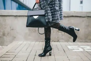 Fashionable person in a fur coat carries a black handbag and wears sheer polka-dot tights with glossy black boots, exuding chic style.