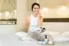 A woman in a white tank top applies face cream while sitting on a bed, smiling. A small mirror and skincare products are on a tray in front of her, creating a cozy, relaxed atmosphere.