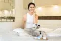 A woman in a white tank top applies face cream while sitting on a bed, smiling. A small mirror and skincare products are on a tray in front of her, creating a cozy, relaxed atmosphere.