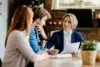 A professional woman in a blue blazer is smiling and discussing a document with a couple at a cozy table, conveying a friendly and collaborative atmosphere.
