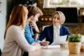 A professional woman in a blue blazer is smiling and discussing a document with a couple at a cozy table, conveying a friendly and collaborative atmosphere.