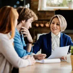 A professional woman in a blue blazer is smiling and discussing a document with a couple at a cozy table, conveying a friendly and collaborative atmosphere.