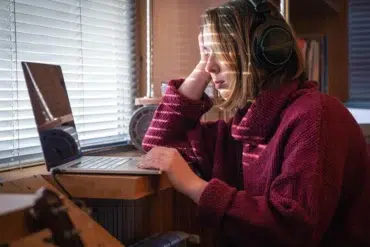 Person in a red sweater and headphones sitting at a wooden desk, looking at a laptop near a window with closed blinds.