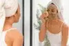 A woman with a towel-wrapped head applies a clay face mask while looking in a mirror. She gently uses a sponge, conveying a calm self-care routine.