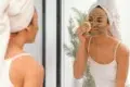 A woman with a towel-wrapped head applies a clay face mask while looking in a mirror. She gently uses a sponge, conveying a calm self-care routine.